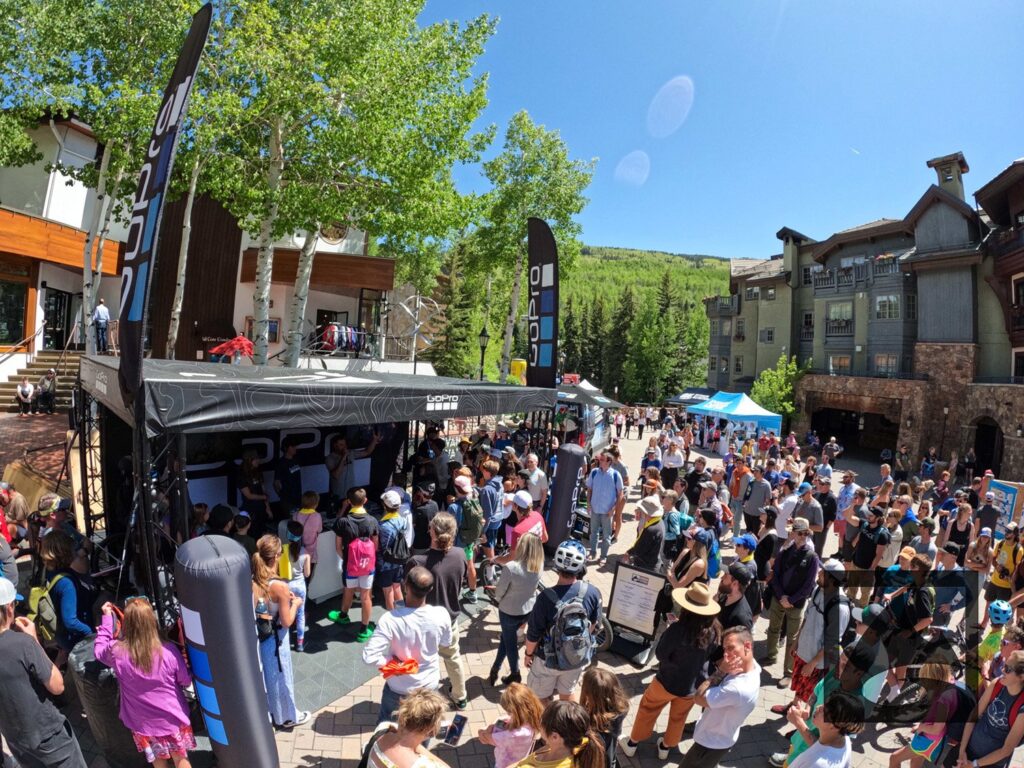 A vibrant outdoor event at the GoPro Mountain Games, with a crowd gathered around a stage set up under tent canopies, flanked by promotional banners.