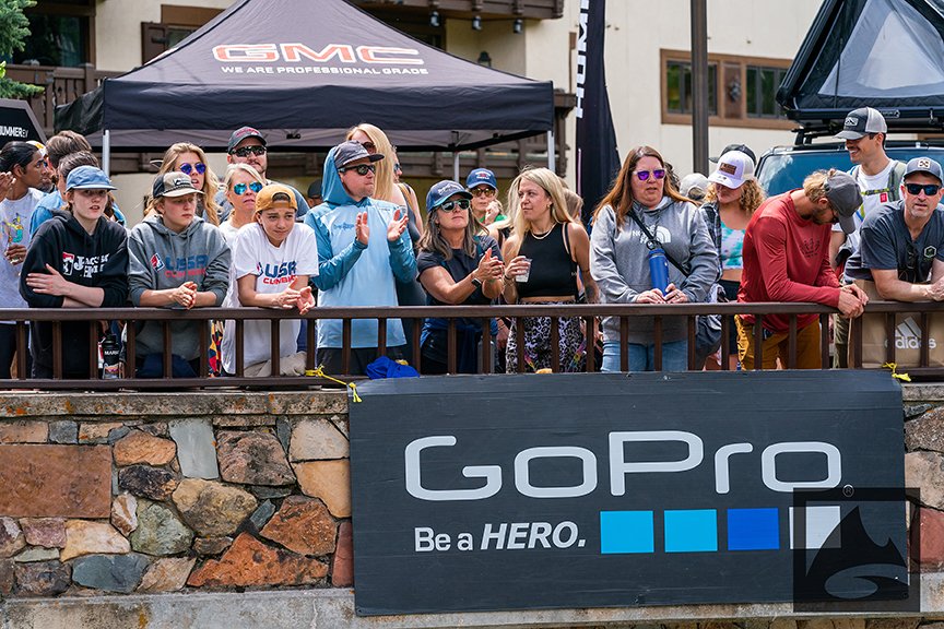 A group of spectators, including adults and children wearing sunglasses, watches an event from behind a GoPro Mountain Games-branded barrier.