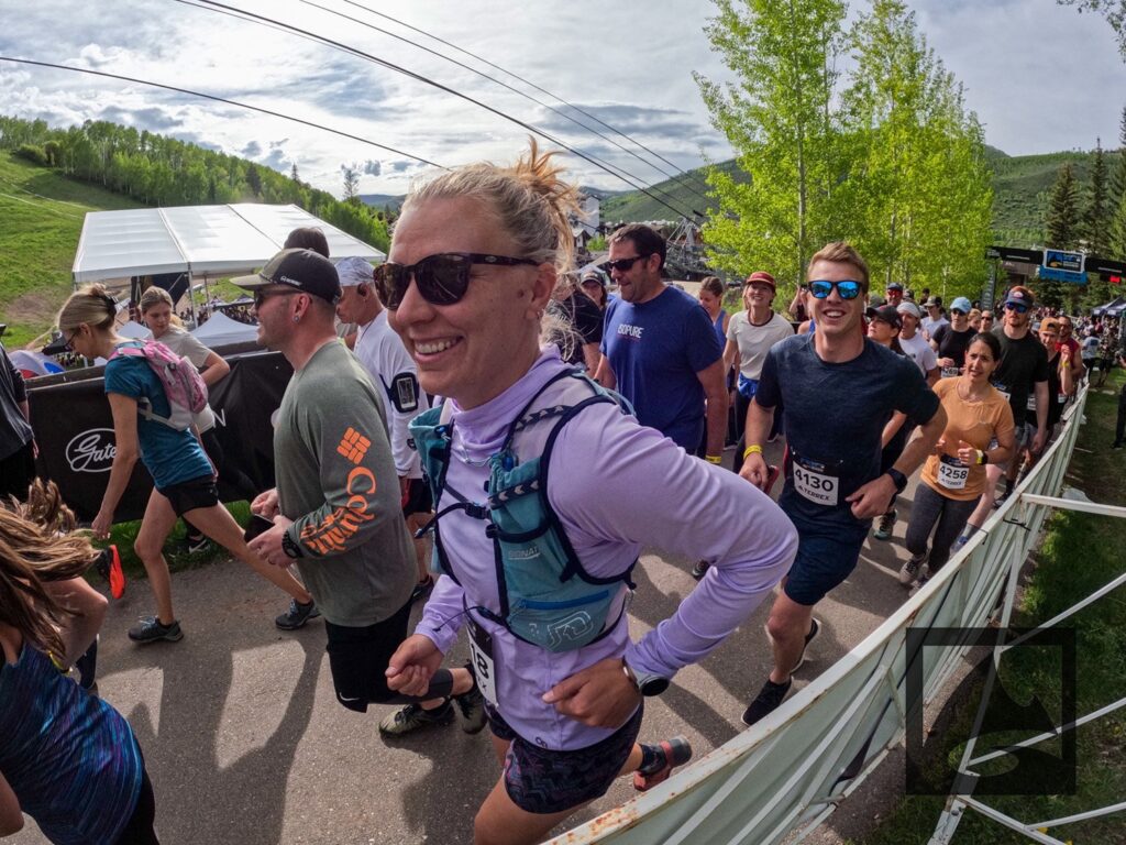 A female runner in sunglasses and a hydration pack smiles at the camera during a crowded race event at the GoPro Mountain Games, with spectators and other runners around her.